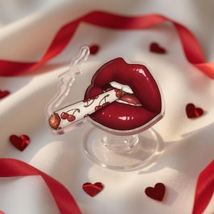 May include: A clear acrylic phone grip featuring a cartoon image of red lips with a cherry-decorated cigarette. The lips are glossy red, and the cigarette has a white background with red cherries. The grip is on a clear stand, with red heart-shaped confetti.