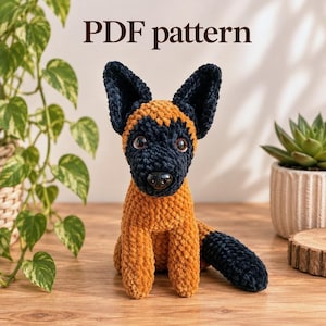 May include: A PDF pattern for a crocheted plush dog toy. The dog is made of soft, textured yarn in shades of golden brown and dark navy blue, featuring large pointed ears, a dark snout, and round brown eyes. The text PDF pattern is visible.