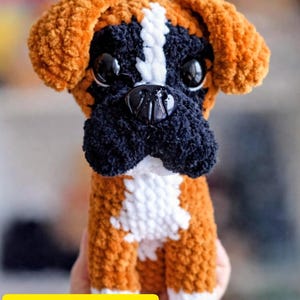 Puede incluir: Un perro bóxer de ganchillo hecho a mano con hilo marrón, negro y blanco. El perro tiene ojos negros grandes y una nariz negra. Las orejas son marrones y el hocico es negro. El texto "PDF pattern" está en un rectángulo amarillo.