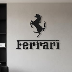 Può includere: Arte da parete in metallo nero con il logo Ferrari, incluso l'emblema del cavallino rampante sopra il nome del marchio. L'opera d'arte è montata su una parete grigio chiaro in un ambiente moderno. Il logo è un simbolo popolare per gli appassionati di auto.