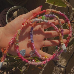 May include: A decorative USB charging cable, hand-crafted with a colourful wrap. The cable is wrapped in pink, red, yellow, and purple threads, decorated with beads and butterfly charms. It has a white USB connector.