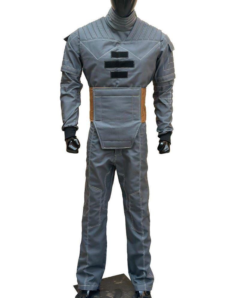May include: A gray jumpsuit with a high collar, featuring a brown belt and black gloves. The suit has white stitching and padded details on the shoulders and chest. The outfit is displayed on a mannequin.