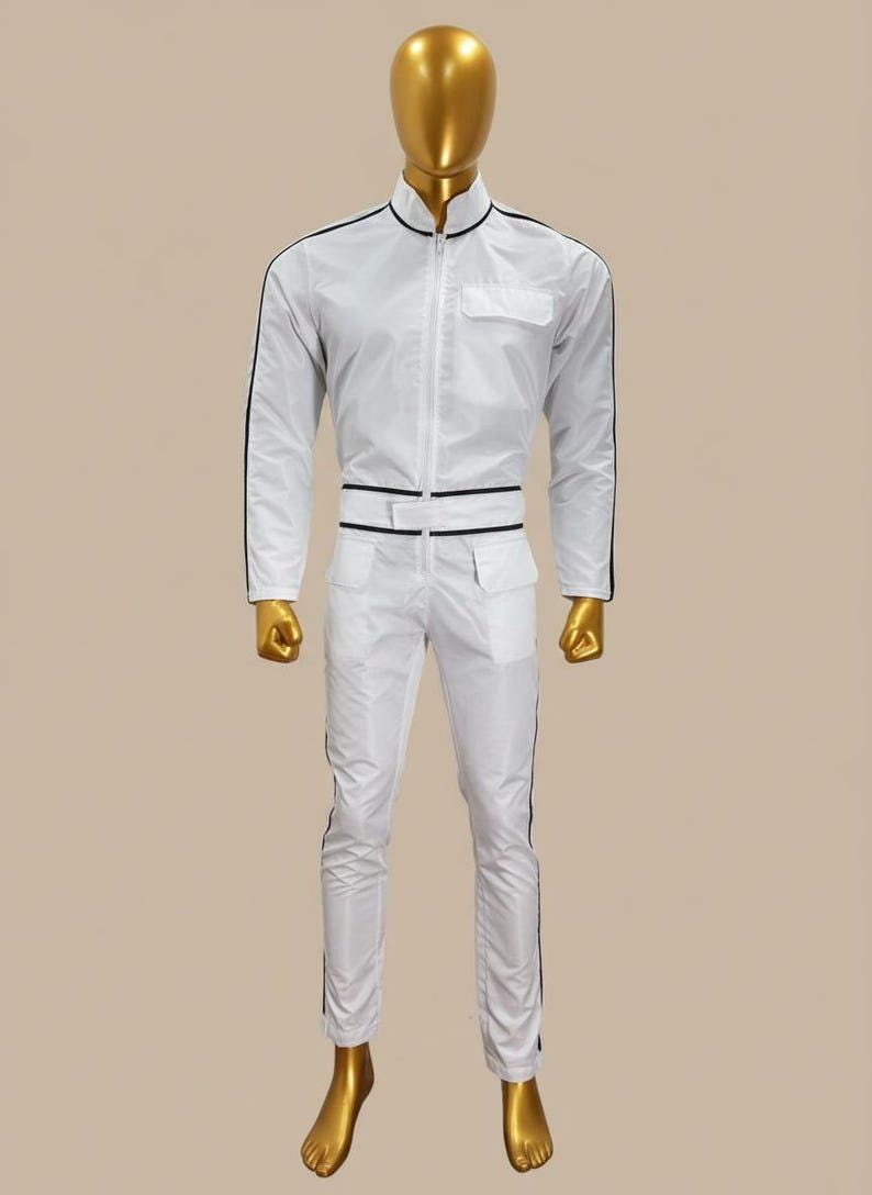 K&ouml;nnte beinhalten: Wei&szlig;er Overall mit schwarzen Zierleisten, Stehkragen und Rei&szlig;verschluss. Das Outfit umfasst eine Brusttasche, zwei H&uuml;fttaschen und ein G&uuml;rteldetail. Die Hose hat schwarze Streifen an den Seiten. Die Schaufensterpuppe ist goldfarben.