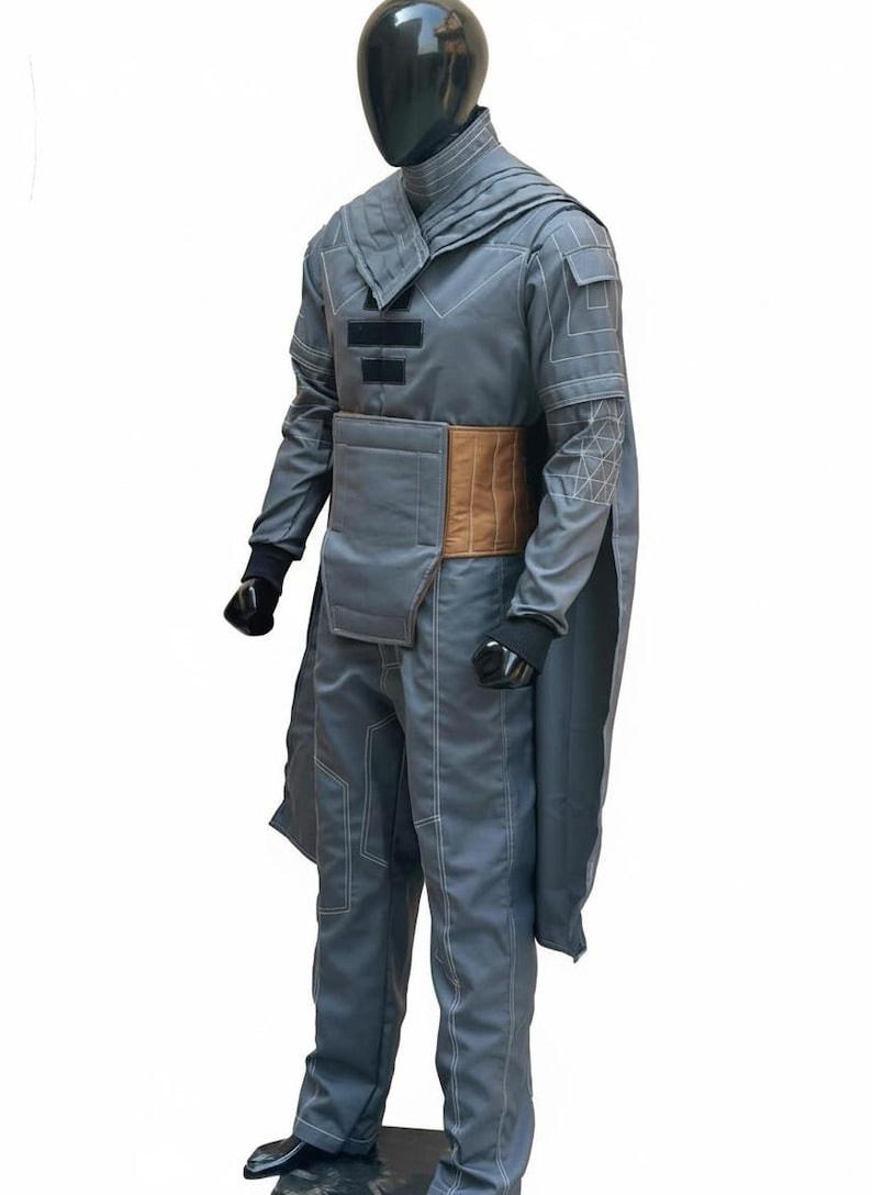May include: A gray costume with a cape, pants, and a jacket. The jacket has a high collar and geometric stitching. A brown belt cinches the waist. The costume is displayed on a mannequin.