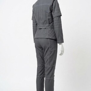 May include: A gray two-piece outfit on a mannequin. The jacket has white stitching and quilted detailing on the back. The pants have pockets and white stitching. The outfit is displayed on a black stand.