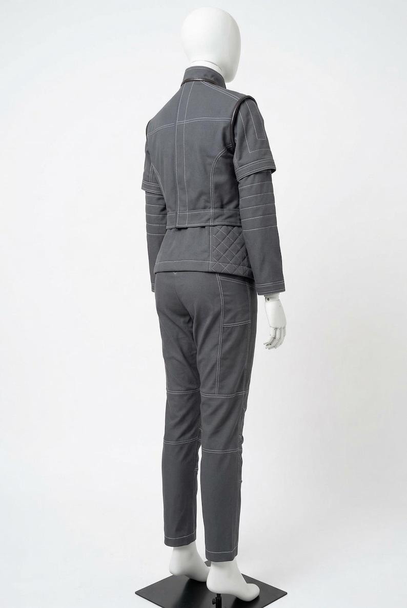 May include: A gray two-piece outfit on a mannequin. The jacket has white stitching and quilted detailing on the back. The pants have pockets and white stitching. The outfit is displayed on a black stand.