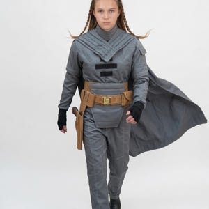 May include: A gray cosplay costume with a cape, belt, and holster. The outfit includes a high-collared top, pants, and a brown belt with a gold buckle. The costume is worn with black gloves and boots.