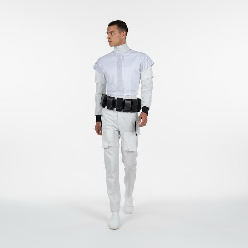 Puede incluir: Un atuendo blanco que consta de un cuello alto, una chaqueta corta, una camisa de manga larga y pantalones. El atuendo incluye un cintur&oacute;n negro con bolsillos y botas blancas. El modelo est&aacute; de pie frente a un fondo blanco.