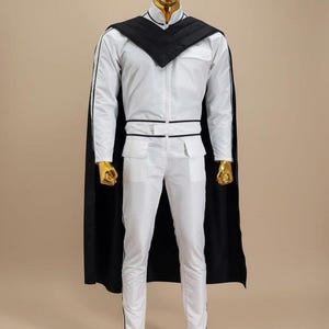 K&ouml;nnte beinhalten: Wei&szlig;er Overall mit schwarzen Zierleisten und passendem schwarzem Umhang. Das Outfit hat einen hohen Kragen, einen Rei&szlig;verschluss vorne und Taschen. Kopf, H&auml;nde und F&uuml;&szlig;e der Schaufensterpuppe sind goldfarben.