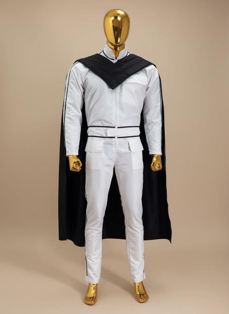 K&ouml;nnte beinhalten: Wei&szlig;er Overall mit schwarzen Zierleisten und passendem schwarzem Umhang. Das Outfit hat einen hohen Kragen, einen Rei&szlig;verschluss vorne und Taschen. Kopf, H&auml;nde und F&uuml;&szlig;e der Schaufensterpuppe sind goldfarben.