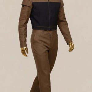 May include: A brown suit displayed on a mannequin. The jacket is black with brown accents and a zipper closure. The pants are brown and straight-legged. The mannequin has a gold head and hands, and is wearing white socks.