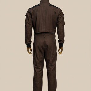 May include: A brown jumpsuit with a high collar and black accents on the shoulders and cuffs. The outfit has a cropped jacket and fitted pants. The mannequin has a gold head and hands.