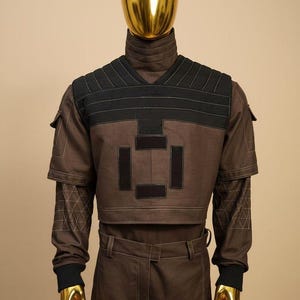 May include: Brown and black futuristic-style outfit. The top has a V-neck with black accents and geometric patches. The long-sleeved shirt has geometric patterns on the sleeves. The outfit includes matching brown pants. The mannequin head and hands are gold.