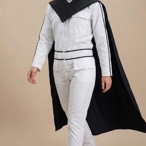 K&ouml;nnte beinhalten: Wei&szlig;er Overall mit schwarzem Besatz und passendem schwarzem Umhang. Der Overall hat lange &Auml;rmel mit schwarzem Streifen, einen Rei&szlig;verschluss vorne und Taschen. Der Umhang ist &uuml;ber den Schultern drapiert. Die Person ist barfu&szlig;.