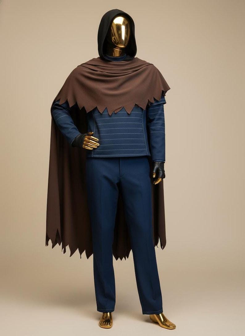 May include: A mannequin dressed in a dark brown cape with a jagged edge, a hooded black top, and navy blue pants. The top has horizontal white stripes. The mannequin's hands and face are gold.