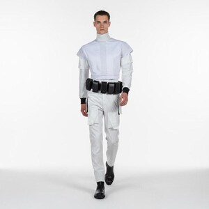 Puede incluir: Un atuendo de estilo futurista en blanco, con una parte superior de cuello alto con l&iacute;neas verticales, pantalones a juego y un cintur&oacute;n negro con m&uacute;ltiples bolsillos. El atuendo incluye detalles en negro en las mangas y los tobillos, y botas negras.
