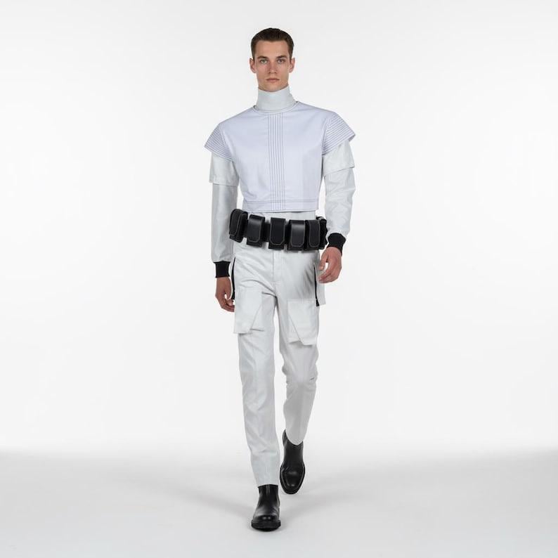 Puede incluir: Un atuendo de estilo futurista en blanco, con una parte superior de cuello alto con l&iacute;neas verticales, pantalones a juego y un cintur&oacute;n negro con m&uacute;ltiples bolsillos. El atuendo incluye detalles en negro en las mangas y los tobillos, y botas negras.