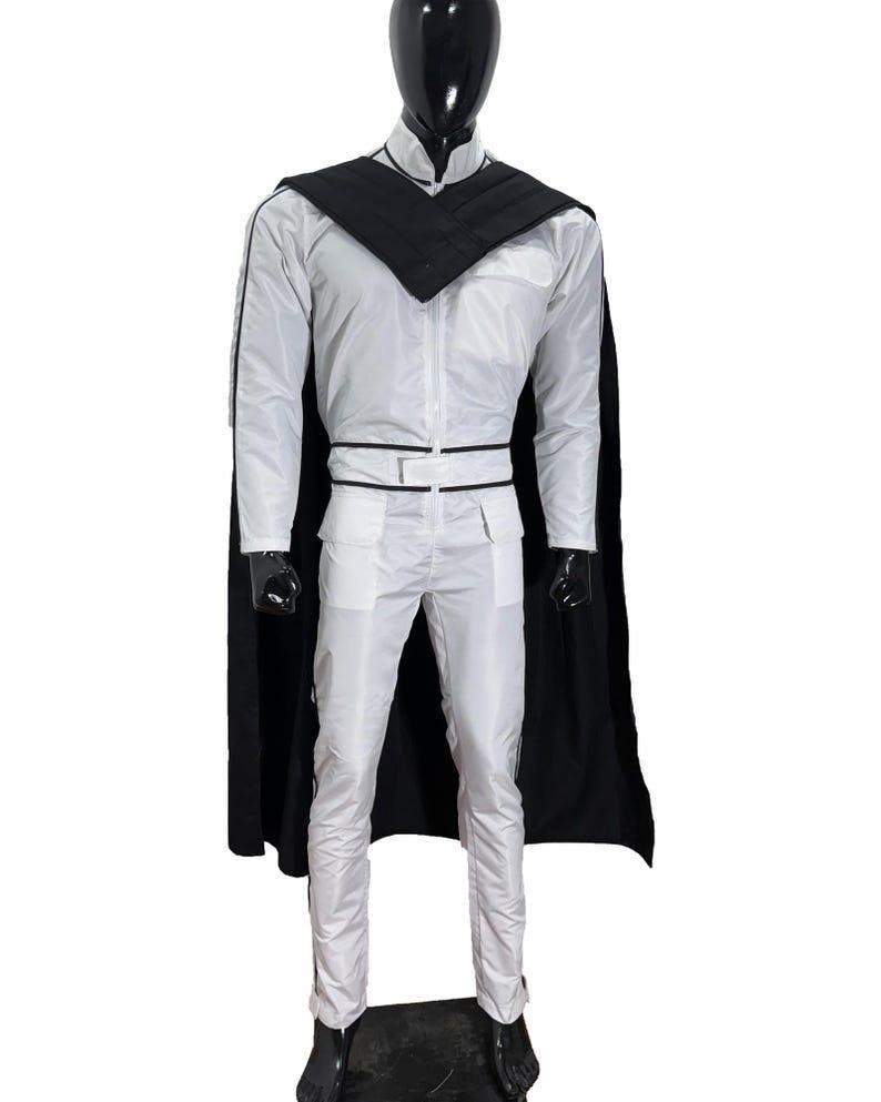 K&ouml;nnte beinhalten: Ein wei&szlig;es Kost&uuml;m mit schwarzem Umhang, Kragen und Besatz. Das Outfit umfasst eine lang&auml;rmlige Jacke, eine Hose und einen G&uuml;rtel. Die Schaufensterpuppe steht auf einem schwarzen Sockel. Das Kost&uuml;m ist wahrscheinlich f&uuml;r Cosplay oder eine Themenveranstaltung.