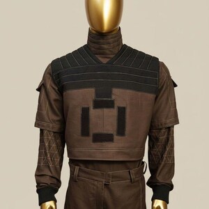 May include: Brown and black two-piece outfit on a mannequin. The top has a high collar and a V-neck with black accents. The sleeves have geometric patterns. The pants are brown with a belt.