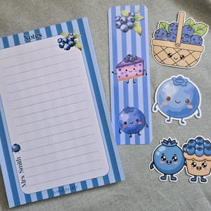 May include: A stationery set featuring a lined notepad with blue stripes and blueberry illustrations, a matching bookmark with cartoon blueberry characters, and three die-cut stickers of a blueberry basket, a blueberry, and a blueberry muffin.