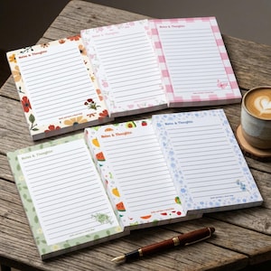 May include: Five rectangular notepads with lined paper, each featuring the header Notes & Thoughts and a unique border design including floral, fruit, and checkered patterns. A brown fountain pen lies horizontally below the notepads.