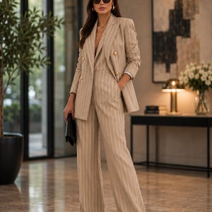 May include: A beige pinstripe suit with a double-breasted blazer and wide-leg trousers. The suit features gold buttons and is paired with a black clutch and sunglasses. The outfit is worn in a well-lit, modern interior setting.
