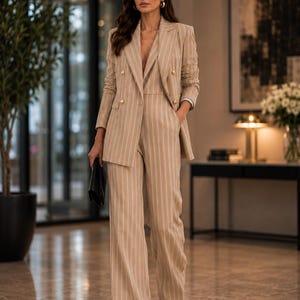 May include: A beige pinstripe suit featuring a double-breasted blazer with gold buttons and wide-leg trousers. The person is wearing sunglasses and carrying a black clutch. The suit's neutral colour makes it suitable for business or formal occasions.