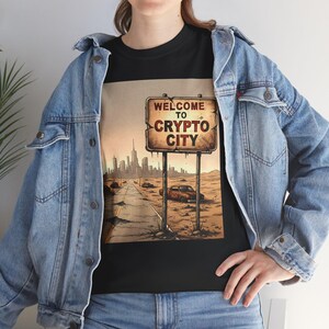 Puede incluir: Camiseta negra con un gráfico de una señal de tráfico que dice "Welcome to Crypto City". La señal está situada en un paisaje desértico con un horizonte urbano. La camiseta se lleva con una chaqueta vaquera.