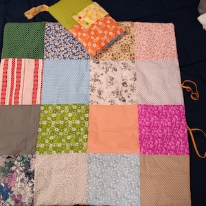 Könnte beinhalten: Eine rechteckige Patchwork-Decke bestehend aus sechzehn Quadraten mit verschiedenen Mustern wie Blumen, Punkten, Streifen und Vichy-Karos in Farben wie Grün, Rot, Blau, Orange, Pink und Braun, inklusive einer angebrachten, gefalteten Stofftasche.