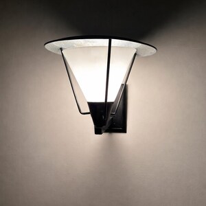 Puede incluir: Aplique de pared moderno con pantalla cónica y parte superior circular. El aplique tiene un marco de metal negro y una pantalla de vidrio esmerilado que emite una luz suave y difusa. Montado en una pared oscura.