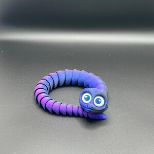 Könnte beinhalten: Eine flexible, bewegliche Spielzeugschlange mit einem Farbverlauf von tiefem Violett zu leuchtendem Blau. Die Schlange hat einen strukturierten, segmentierten Körper und einen abgerundeten Kopf mit großen, auffälligen blau-weißen Comic-Augen.