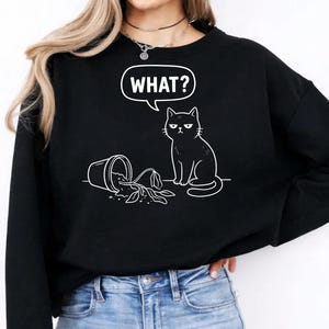 Könnte beinhalten: Schwarzes Sweatshirt mit einer weißen Grafik einer Katze neben einem umgestürzten Blumentopf. Die Katze hat einen unbeeindruckten Gesichtsausdruck, und eine Sprechblase darüber sagt "WHAT?"