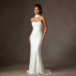 May include: A floor-length white gown featuring a strapless sweetheart neckline, a fitted bodice with subtle ruching, and a high-neck collar attachment. The fabric displays a tonal floral pattern, and the skirt flows into a slight train.