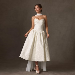 May include: A white strapless midi dress featuring a subtle tonal floral jacquard pattern, a fitted bodice with a sweetheart neckline, and a full, pleated skirt. The garment includes a matching sheer white neck scarf draped around the shoulders.