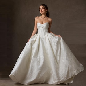 May include: A strapless ivory ball gown featuring a structured corset bodice with subtle pleating and a voluminous floor-length skirt crafted from a textured jacquard fabric with a tonal floral pattern throughout the entire garment.