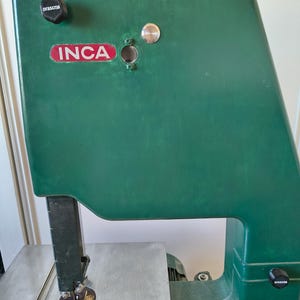 Peut inclure: Une scie à ruban INCA verte dotée d'une table de travail en métal, d'un guide-lame vertical et de boutons de réglage noirs marqués orascna. La machine présente un logo INCA rouge et blanc sur le capot supérieur et un bouton circulaire argenté.