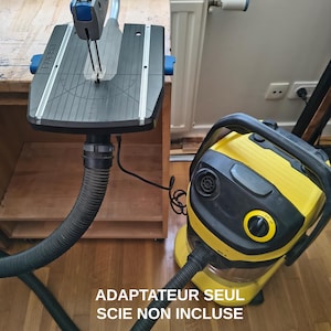 Peut inclure: Un adaptateur de scie à chantourner Dremel avec une surface de travail noire et un bras gris, relié par un tuyau noir à un aspirateur eau et poussière jaune et noir. Le texte ADAPTATEUR SEUL SCIE NON INCLUSE ADAPTER ONLY SAW NOT INCLUDED est présent.