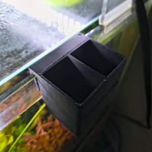 Op de afbeelding: Een zwart, rechthoekig aquariumaccessoire met drie compartimenten. Het item is bevestigd aan de zijkant van een glazen aquarium, met waterplanten en water zichtbaar op de achtergrond. Het accessoire lijkt 3D-geprint te zijn.