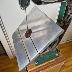 May include: A metal bandsaw featuring a large, flat, silver-colored work table with a central slot and a circular dark red insert. The vertical blade passes through the center of the table, which is mounted on a dark green frame and base.