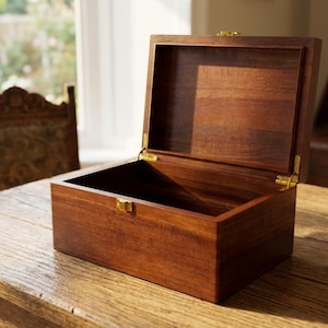 May include: A wooden box with a hinged lid, open to reveal the interior. The box is a rich brown color, with brass-colored hardware. It sits on a wooden table, with a window and chair in the background. The box is likely used for storage or display.