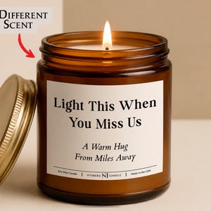 May include: A lit soy wax candle in an amber glass jar with a gold metal lid. The white label features the black text: Light This When You Miss Us, A Warm Hug From Miles Away, Soy Wax Candle, AYSBERG NJ CANDLE, Made in the USA. A red arrow points to the text: 8 Different Scent.