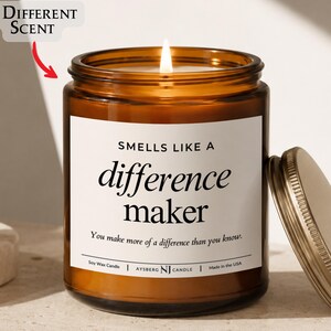 May include: A lit soy wax candle in an amber glass jar with a metal lid. The white label reads: "SMELLS LIKE A difference maker. You make more of a difference than you know." Text above the candle indicates "8 DIFFERENT SCENT" with a red arrow.
