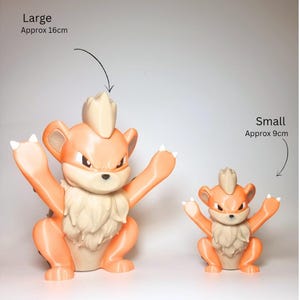 May include: Two orange and cream-colored creature figurines with pointed ears and tufted fur. The larger figure is labeled Large Approx 16cm and the smaller figure is labeled Small Approx 9cm. Both figures have raised arms and a determined expression.