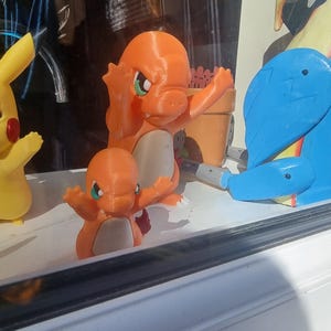 May include: Two orange Charmander figures with cream-colored bellies and green eyes stand in a row. To the left is a yellow Pikachu figure, and to the right is a blue and yellow bird-like figure. The figures are positioned on a white ledge.