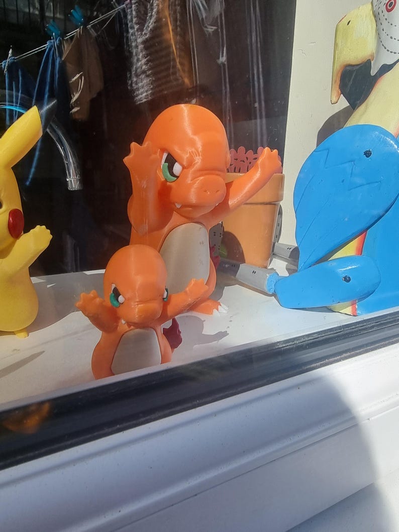 May include: Two orange Charmander figures with cream-colored bellies and green eyes stand in a row. To the left is a yellow Pikachu figure, and to the right is a blue and yellow bird-like figure. The figures are positioned on a white ledge.