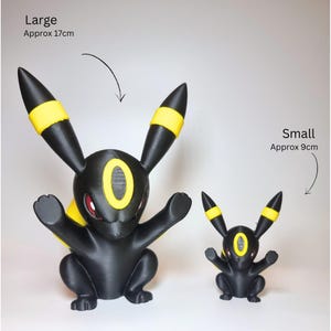 May include: Two black and yellow Umbreon figures with red eyes. The large figure is labeled Large Approx 17cm and the small figure is labeled Small Approx 9cm. Both figures have long ears with yellow bands and a yellow oval marking on the forehead.