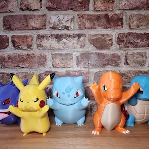 May include: A collection of five colorful plastic character figurines including a purple Gengar, a yellow Pikachu, a light blue Bulbasaur, an orange Charmander, and a blue Squirtle, all standing in a row on a wooden surface.