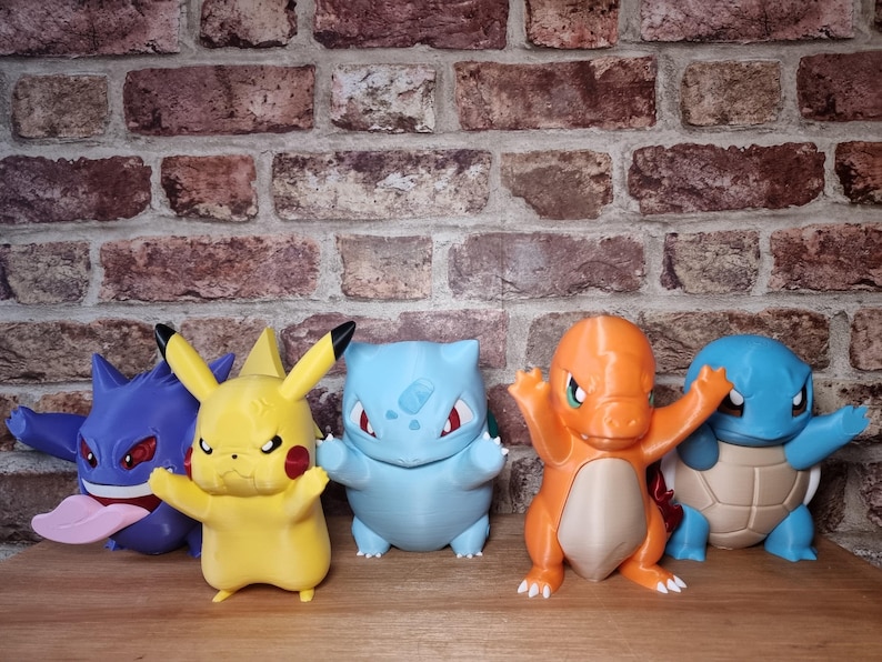 May include: A collection of five colorful plastic character figurines including a purple Gengar, a yellow Pikachu, a light blue Bulbasaur, an orange Charmander, and a blue Squirtle, all standing in a row on a wooden surface.