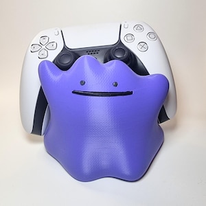 May include: A purple, amorphous, blob-shaped controller stand featuring two small black dot eyes and a thin black horizontal line for a mouth. The stand is holding a white and black wireless video game controller with standard buttons and joysticks.