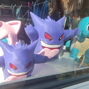 May include: Two purple Gengar figures with pointed ears, red eyes, and wide grins showing white teeth and pink tongues. To the left is a light pink figure with large ears, and to the right is a light blue figure with a tan shell on its back.